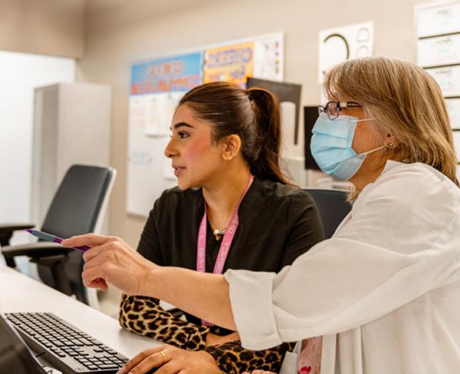 Two women, one masked and in a doctor's coat, stare at a computer monitor.
