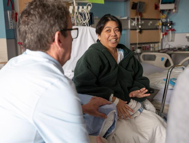 Female patient on a hospital bed looking at a health care provider who has sat down across from her