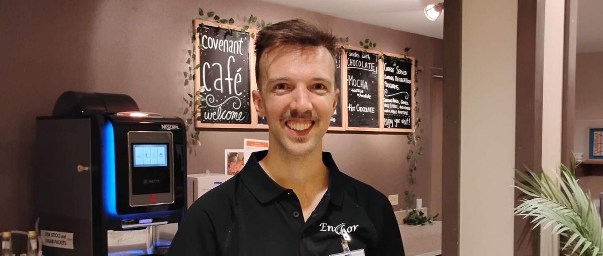 Covenant Care recreation assistant stands in front of a coffee machine and a welcome sign for the Covenant Cafe