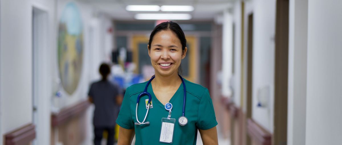 Makayla Mantla, an undergraduate nursing employee at St. Joseph's General Hospital in Vegreville, stands smiling in a brightly lit hospital corridor wearing green scrubs and a stethoscope around her neck.