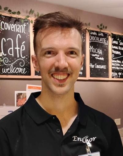 Covenant Care recreation assistant stands in front of a coffee machine and a welcome sign for the Covenant Cafe