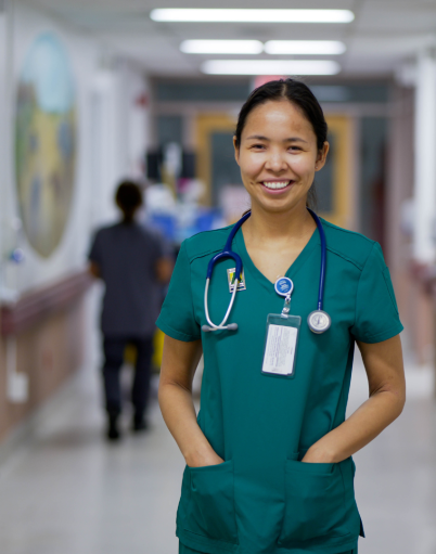 Makayla Mantla, an undergraduate nursing employee at St. Joseph's General Hospital in Vegreville, stands smiling in a brightly lit hospital corridor wearing green scrubs and a stethoscope around her neck.