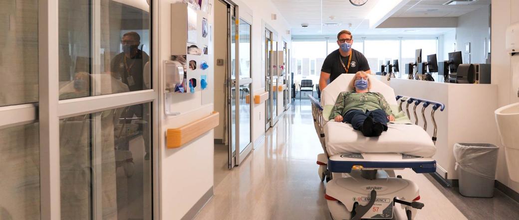 A healthcare worker transports a patient on a gurney down a hospital hallway.