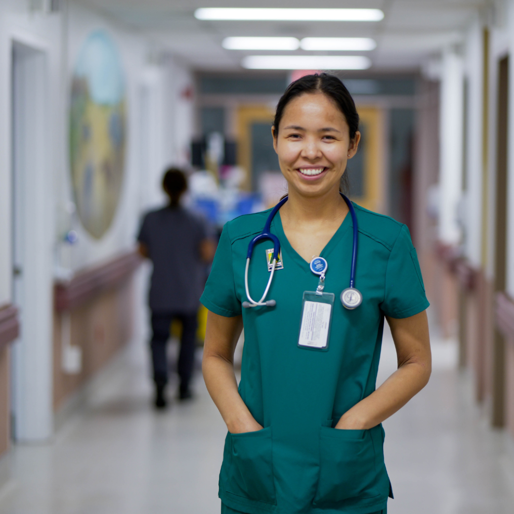 Makayla Mantla, an undergraduate nursing employee at St. Joseph's General Hospital in Vegreville, stands smiling in a brightly lit hospital corridor wearing green scrubs and a stethoscope around her neck.