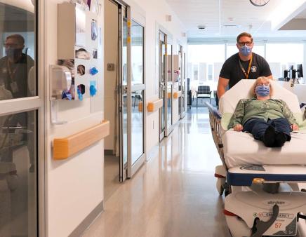 A healthcare worker transports a patient on a gurney down a hospital hallway.